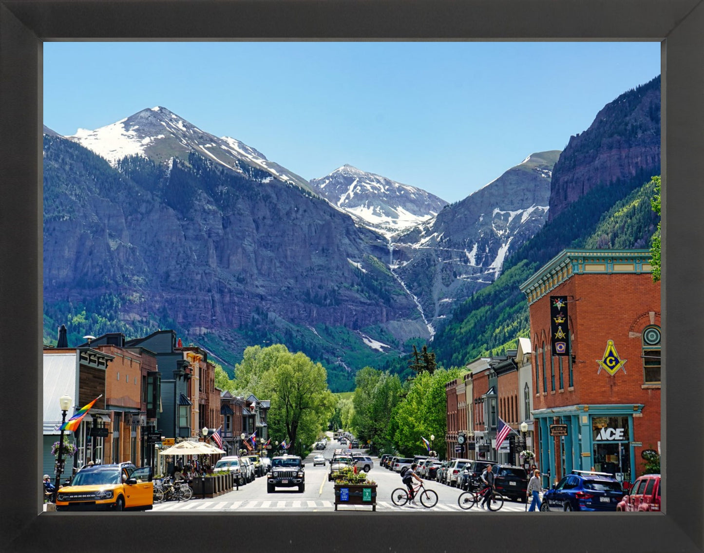 A framed photograph of a small town with mountains in the background, featuring a street with parked cars and buildings.