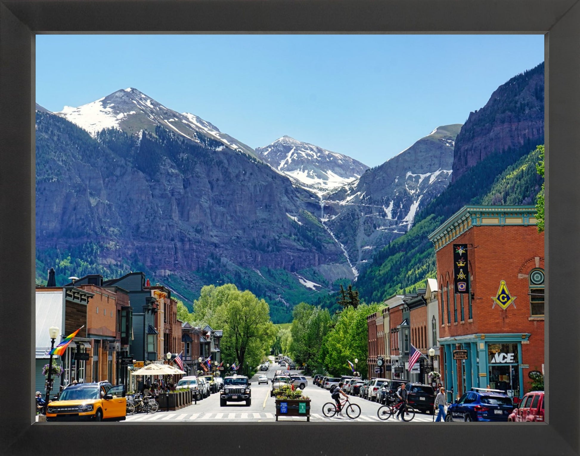 A framed photograph of a small town with mountains in the background, featuring a street with parked cars and buildings.