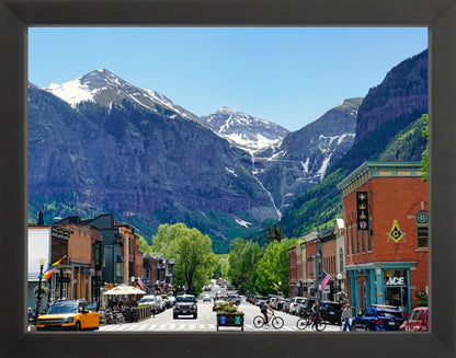 A framed photograph of a small town with mountains in the background, featuring a street with parked cars and buildings.