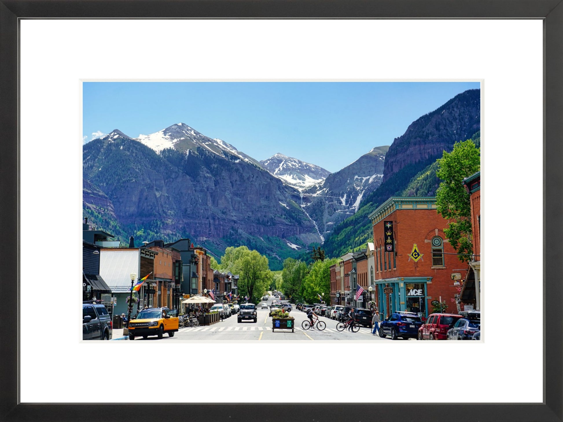 A framed photograph of a small town with mountains in the background, featuring a street with parked cars and buildings.